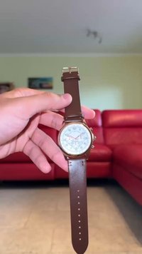 Ruskea nahkainen rannekello, jossa on pyöreä valkoinen kellotaulu, arabialaiset numerot ja kultaiset yksityiskohdat. Varustettu solki-kiinnityksellä ja koristeellisilla kuvioilla kellotaulussa.