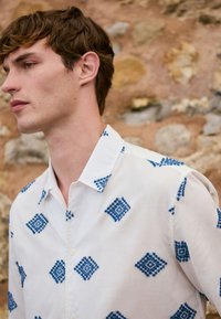 Young man with short brown hair wearing a white shirt with blue geometric patterns, standing against a textured stone wall background.