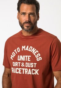 Red short-sleeve T-shirt made of cotton, featuring large white text: "MOTO MADNESS UNITE DIRT & DUST RACETRACK." Simple crew neck design.