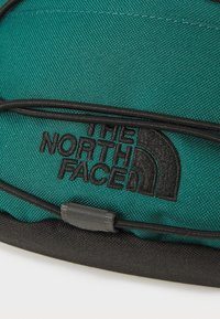 Teal színű szövet hátizsák fekete részletekkel, a hátsó oldalon egy nagy hímzett logóval, melyen a "THE NORTH FACE" felirat szerepel, valamint az elején bungee zsinóros részlet található.