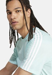 Giovane con dreadlock lunghi che indossa una maglietta Adidas azzurro chiaro con strisce bianche, che guarda leggermente di lato su uno sfondo neutro.