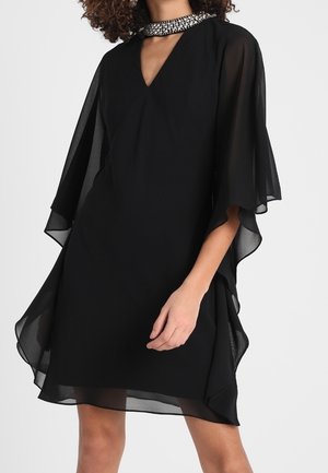 Czarna przezroczysta sukienka z szyfonu z luźnymi rękawami w formie peleryny i koralikowym, wysokim kołnierzem, noszona przez modelkę o kręconych włosach.