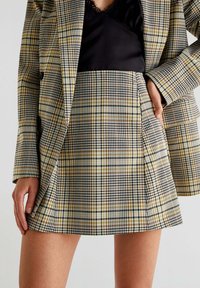 Ruteforstykke i gul, sort og brun med en plisseret design og flæser. Sammen med en sort top skaber det et lag-på-lag look.
