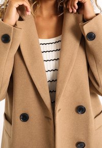Mujer ajustando el cuello de un abrigo de color camel de doble botonadura con botones negros, vistiendo una camiseta blanca de rayas horizontales en negro.
