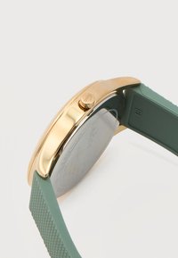 Θήκη ρολογιού από χρυσό με υφασμένο πράσινο λουράκι από σιλικόνη, που δείχνει το πλάγιο κουμπί και την πίσω πλευρά από ανοξείδωτο ατσάλι.