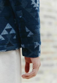 Rukáv modrej kockovanej bundy s geometrickými tvarmi, jemnou tkaninou a jedným čiernym gombíkom na manžete.