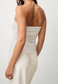 Axelbandslös vit topp i mjukt tyg med rynkning på sidan; kombinerad med en slät vit kjol. Synligt ärrdetalj på ryggen.