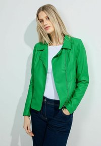 Veste de moto en simili cuir vert avec un col cranté, fermeture éclair à l'avant et deux poches latérales, portée avec un haut blanc et un jean foncé.