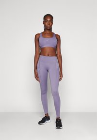 Világos lila sportmelltartó és hozzá illő, magas derekú leggings cipzáras zsebekkel, nedvességelvezető anyagból készült. Fekete sportcipők fehér kiegészítőkkel.