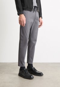 Grå bomullsbyxor med avslappnad passform, kombinerade med svarta snörskor och ribbade svarta strumpor, mot en neutral bakgrund.