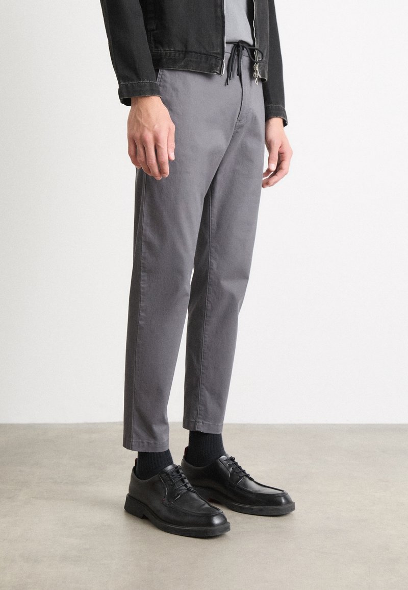 Grå bomullsbyxor med avslappnad passform, kombinerade med svarta snörskor och ribbade svarta strumpor, mot en neutral bakgrund.