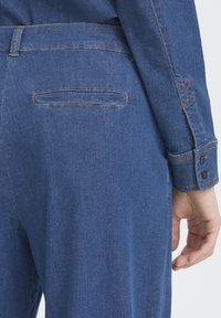 Chemise en denim bleu foncé à texture lisse, dotée d'un poignet à boutons et d'une poche plaquée à l'arrière, agrémentée de surpiqûres contrastantes.