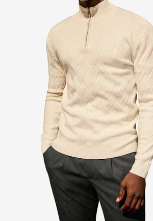 Homme portant un pull beige texturé à fermeture éclair quart avec un motif chevron et un pantalon gris foncé, mains dans les poches.