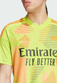 Vihreä urheilu-paita oransseilla korostuksilla. Varustettu verkkokudoksella, rohkealla "Emirates Fly Better" -logolla ja abstrakteilla kuvioilla. Adidas-logomerkintä näkyvissä.
