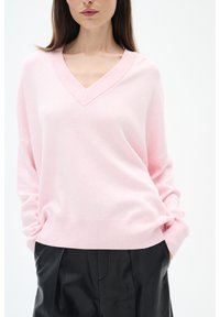 Gaiši rozā V-veida kakla džemperis, izgatavots no mīksta auduma. Tam ir nolaižamas plecu šuves un ribota apakša. Kombinēts ar melniem augstās jostas vietas biksēm.