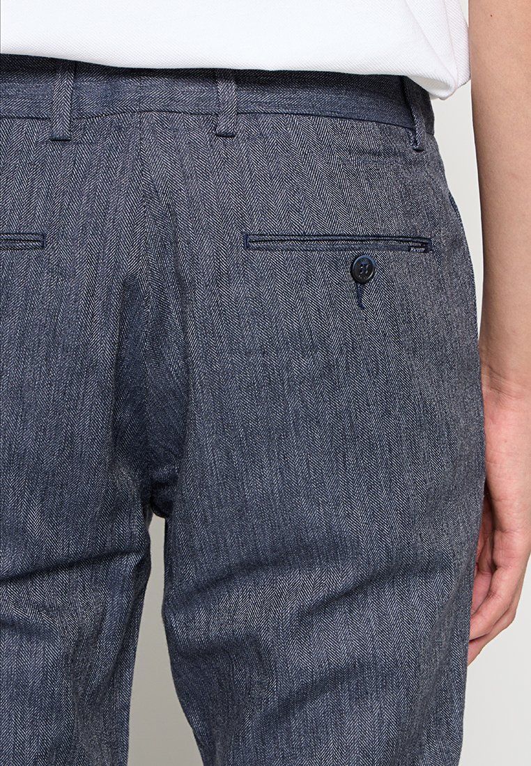 Grau karierte Hosen mit einer knopfverschließbaren Gesäßtasche, die eine strukturierte Webart und eine taillierte Passform aufweisen. Sichtbarer Bund und Gürtelschlaufen.