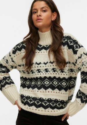 Maglione lavorato a maglia con colletto alto, base crema con motivi geometrici neri e polsini a coste. Texture morbida con dettagli a intaglio.