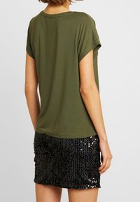 Camiseta oversized color verde oliva con mangas enrolladas, combinada con una falda corta negra de lentejuelas, que muestra una superficie brillante y un detalle texturizado.