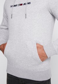 Szary hoodie wykonany z miękkiego materiału, z kieszenią z przodu, ściągaczami na mankietach i logo w kolorze granatowym i czerwonym na piersi.