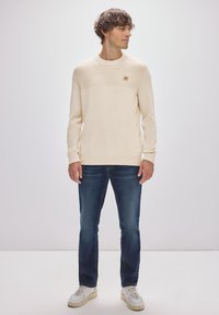 Krämfärgad stickad tröja med horisontell ribbad detalj upptill, tillsammans med mörkblå jeans och vita sneakers.