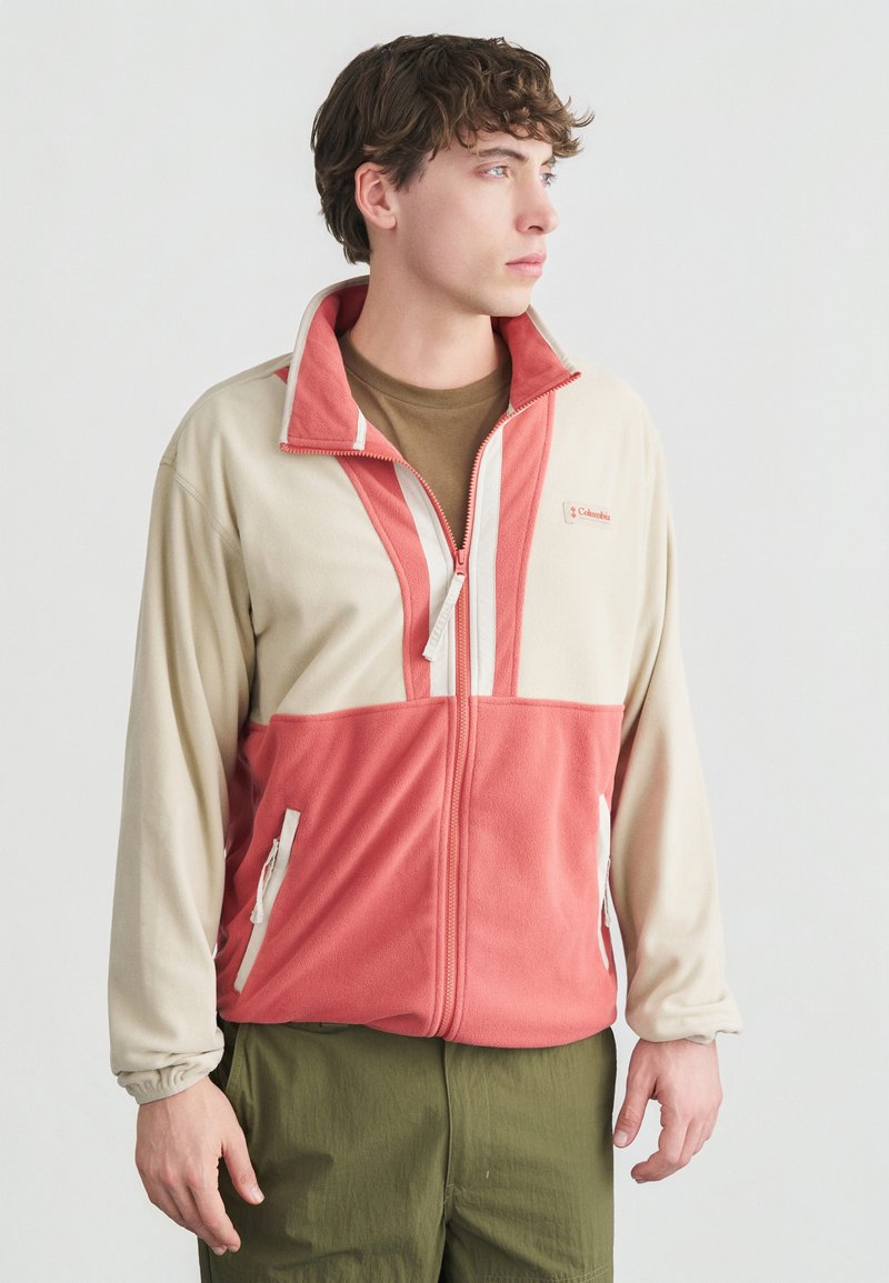 Νεαρός άνδρας με κοντά καστανά μαλλιά που φοράει μπεζ και κοραλλί φλις ζακέτα με φερμουάρ και ελιάς πράσινο παντελόνι, κοιτάζοντας προς τα δεξιά σε απλό φόντο.