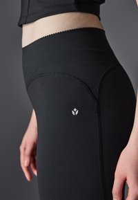 Leggings noirs avec une texture lisse, taille haute et bordure décorative en forme de vagues ; présentent un petit logo blanc sur la hanche.