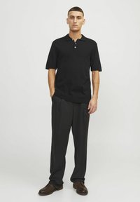 Černé polo tričko s krátkým rukávem vyrobené z pleteného materiálu, se stojáčkem a zapínáním na tři knoflíky, kombinované s tmavými, volnými kalhotami a hnědými botami.