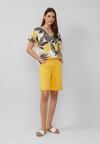 Korte mouwen, patroontop in wit, zwart, en gele bladeren; gecombineerd met knie-lange gele shorts; witte slip-on sandalen.