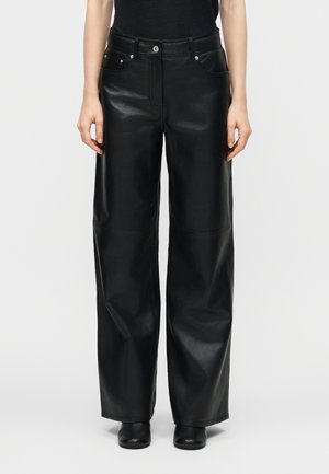 Personne portant un pantalon en cuir noir à jambes larges et taille haute avec des poches avant et des chaussures noires, sur un fond clair uni.