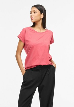 Kvinde med sort, glat hår iført en koralfarvet T-shirt med korte ærmer og sorte bukser, stående med hænderne i lommerne og kigger mod venstre.