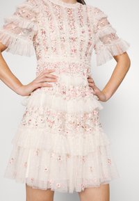 Robe blanche transparente avec broderie florale rose, volants à étages et manches courtes bouffantes, portée avec les mains sur les hanches.