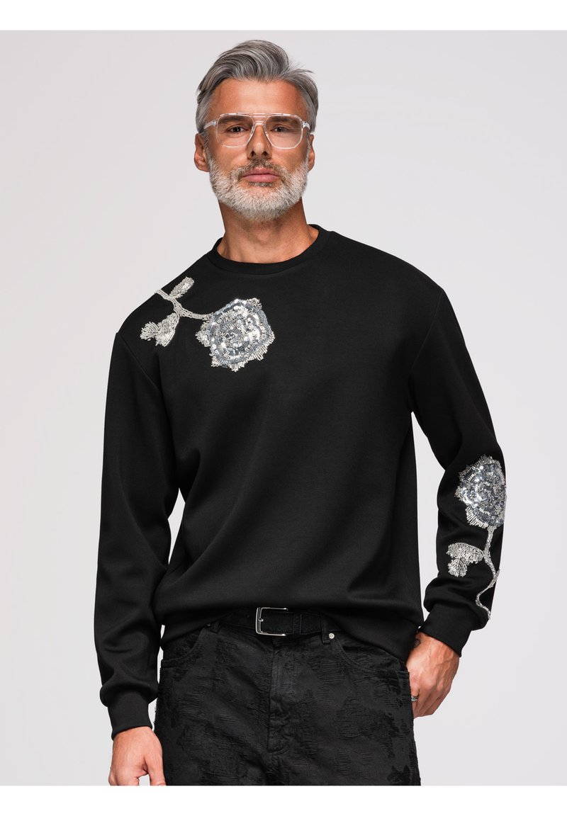 Homme aux cheveux gris et à la barbe portant des lunettes transparentes et un sweat-shirt noir avec des motifs floraux en sequins argentés sur l’épaule et la manche.