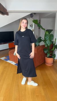 Νεαρή γυναίκα με μαύρο μίντι φόρεμα και λευκά αθλητικά παπούτσια στέκεται χαμογελαστή στο σαλόνι με πορτοκαλί καναπέ και μεγάλο φυτεμένο φυτό.