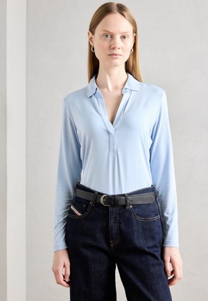 Camisa polo de manga larga azul claro con cuello y una parte de botonadura, combinada con jeans de mezclilla azul oscuro y un cinturón negro.