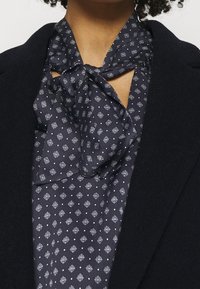 La blusa navy presenta un design con colletto a cravatta e un motivo geometrico bianco. Il tessuto sembra liscio, abbinato a un giubbotto esterno nero.