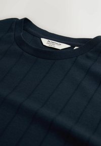 Jūras zila šaura T-krekls ar ribotu struktūru un horizontālām svītrām, ar apaļu kakla izgriezumu un etiķeti, kas rāda zīmola informāciju.