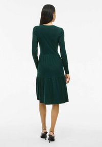 Robe verte foncée à manches longues avec un corsage ajusté et une jupe en couches. Fabriquée en tissu doux, elle présente un col rond et une texture lisse.