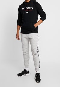 Svart hoodie med vit och röd "Hollister" text. Ljusgrå sweatpants med svarta sidostriper. Svarta sportskor.