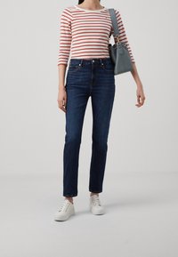 Mujer vestida con una camiseta de rayas rojas y blancas de manga larga, jeans de mezclilla oscura, zapatillas blancas y llevando un bolso azul claro al hombro.
