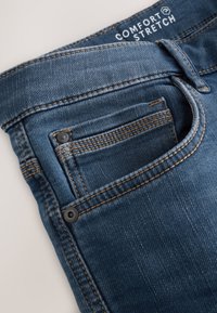 Primo piano della tasca di un jeans in denim blu con cuciture visibili, rivetti e etichetta "Comfort Stretch" all'interno della cintura.