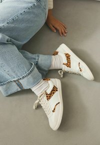 White sneakers featuring leopard print accents on the sides, paired with rolled-up light blue jeans and white socks, on a smooth gray surface.