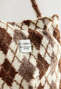 Pruuni ja kreemja ruudulise pehme padja peal on nähtav silt, millel on kirjaga "WILLIAMSBURG BEDFORD AVENUE NEW YORK."
