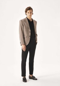 Hellgrauer, strukturierter Blazer mit schwarzem Poloshirt, schlanken schwarzen Hosen und schwarzen Loafers. Steht vor einem schlichten weißen Hintergrund.