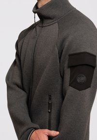 Szara kurtka zapinana na zamek z wysokim kołnierzem, wyposażona w czarną łatę na rękawie oraz boczne kieszenie zapinane na zamek. Wykonana z teksturowanego materiału o miękkim wyglądzie.