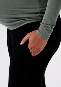 Langarm, aansluitend top van zachte groene stof, gecombineerd met zwarte, elastische broek met zijzakken, waarbij de hand in de zak rust.