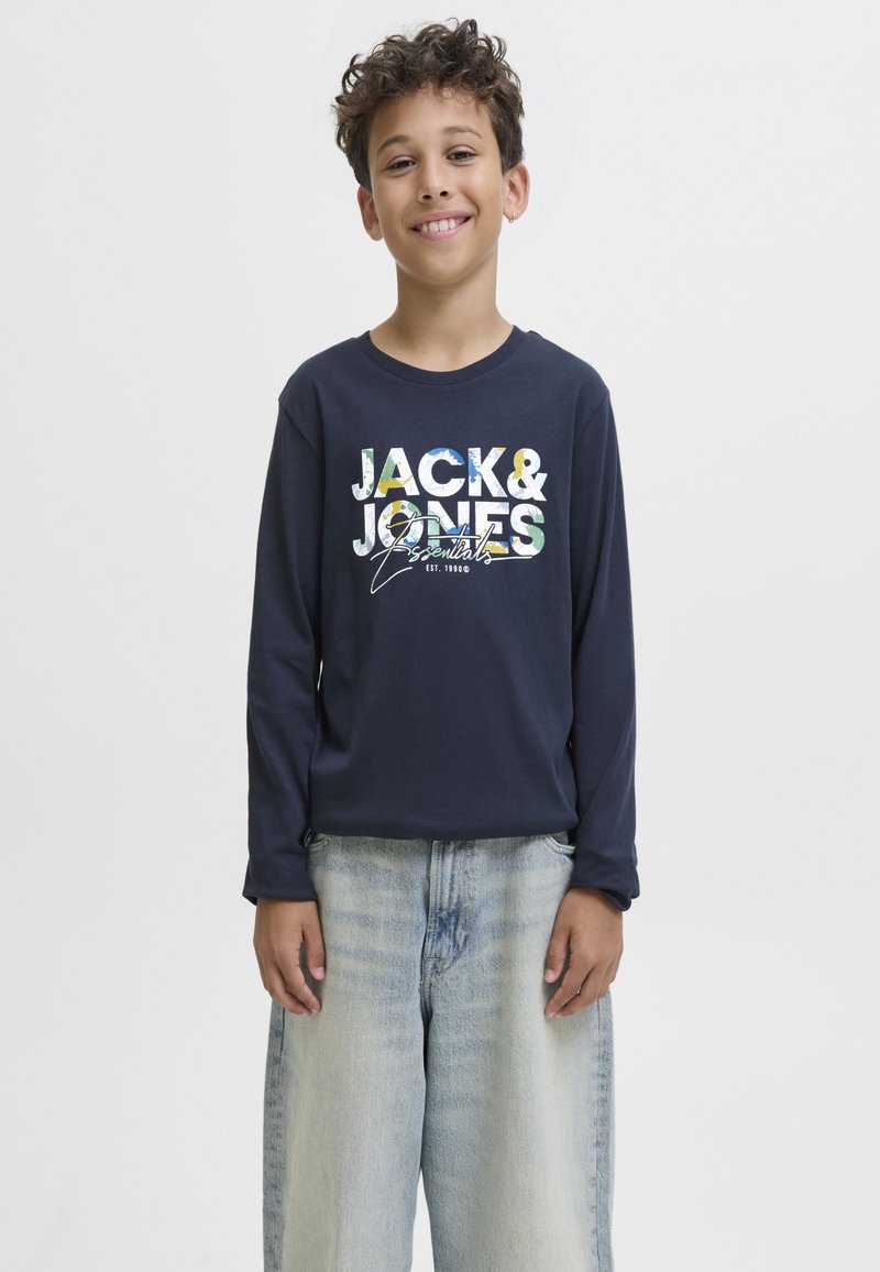 Lächelnder Junge mit lockigem Haar, der ein dunkelblaues Langarmshirt mit mehrfarbigem Jack & Jones-Logo und hellblaue, weite Jeans trägt.