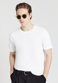 Giovane uomo che indossa una camicia bianca a maniche corte, pantaloni neri con una cintura e occhiali da sole traslucidi marroni, posando con le mani nelle tasche.