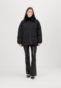 Black puffer jacket with a faux fur collar, front zipper, and boxy fit. Worn with fitted black pants, standing against a plain background.
