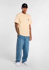 Beige cotton t-shirt with a small chest pocket, paired with light blue baggy denim jeans and white sneakers. Black cap completes the outfit.