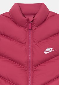 Roza puhovka z visokim ovratnikom in cikcakastim šivom. Ima sprednji zadrgo in bel Nike logotip na levi prsni strani. Gladka tekstura.